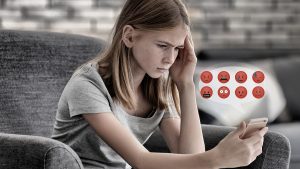 Ein Bild von einem Mädchen das auf ein Smartphone starrt. Ihr Gesichtausdruck ist eher verzweifelt, neben Ihr sind auch traurige und böse Emojis abgebildet.